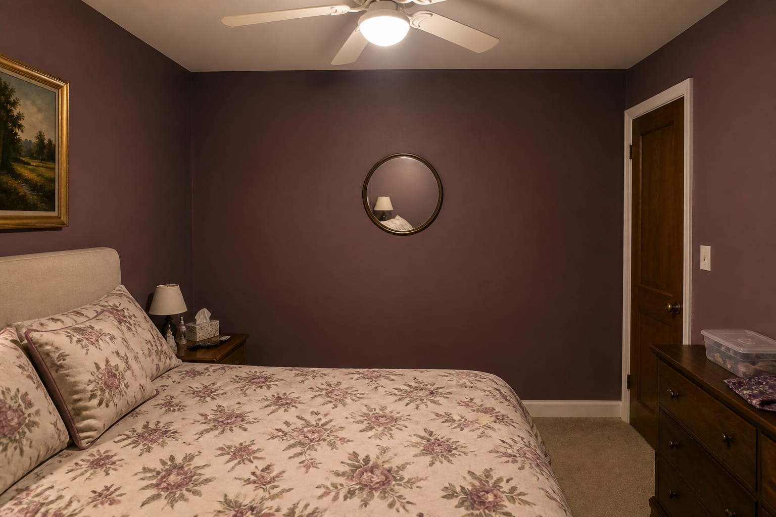 Windowless bedroom with deep plum walls, a small round mirror away from the light, floral bedding, and a cold ceiling-fan light.
