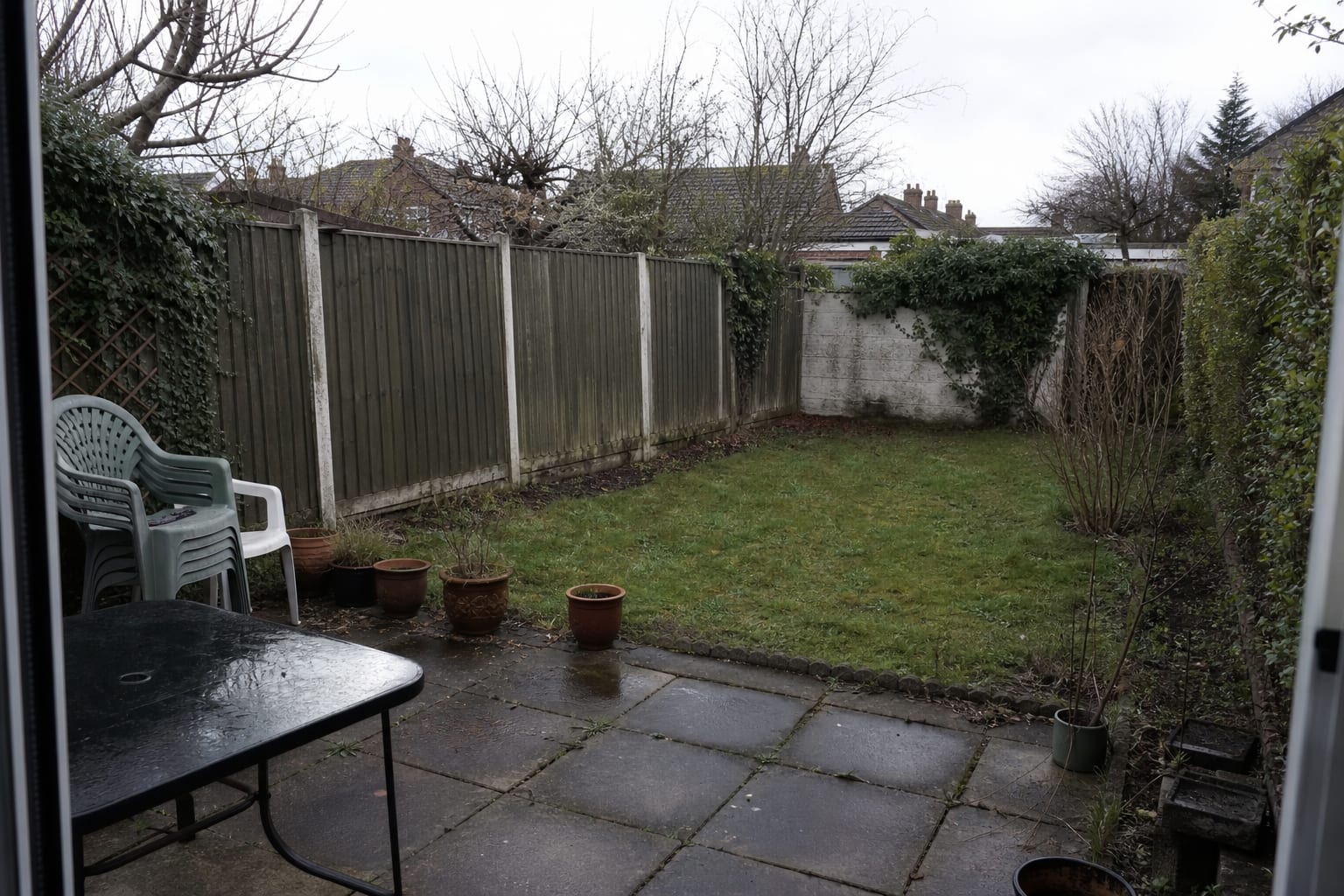 underused winter backyard with bare lawn, stacked chairs, dark patio corner, empty planters, and no clear place to sit