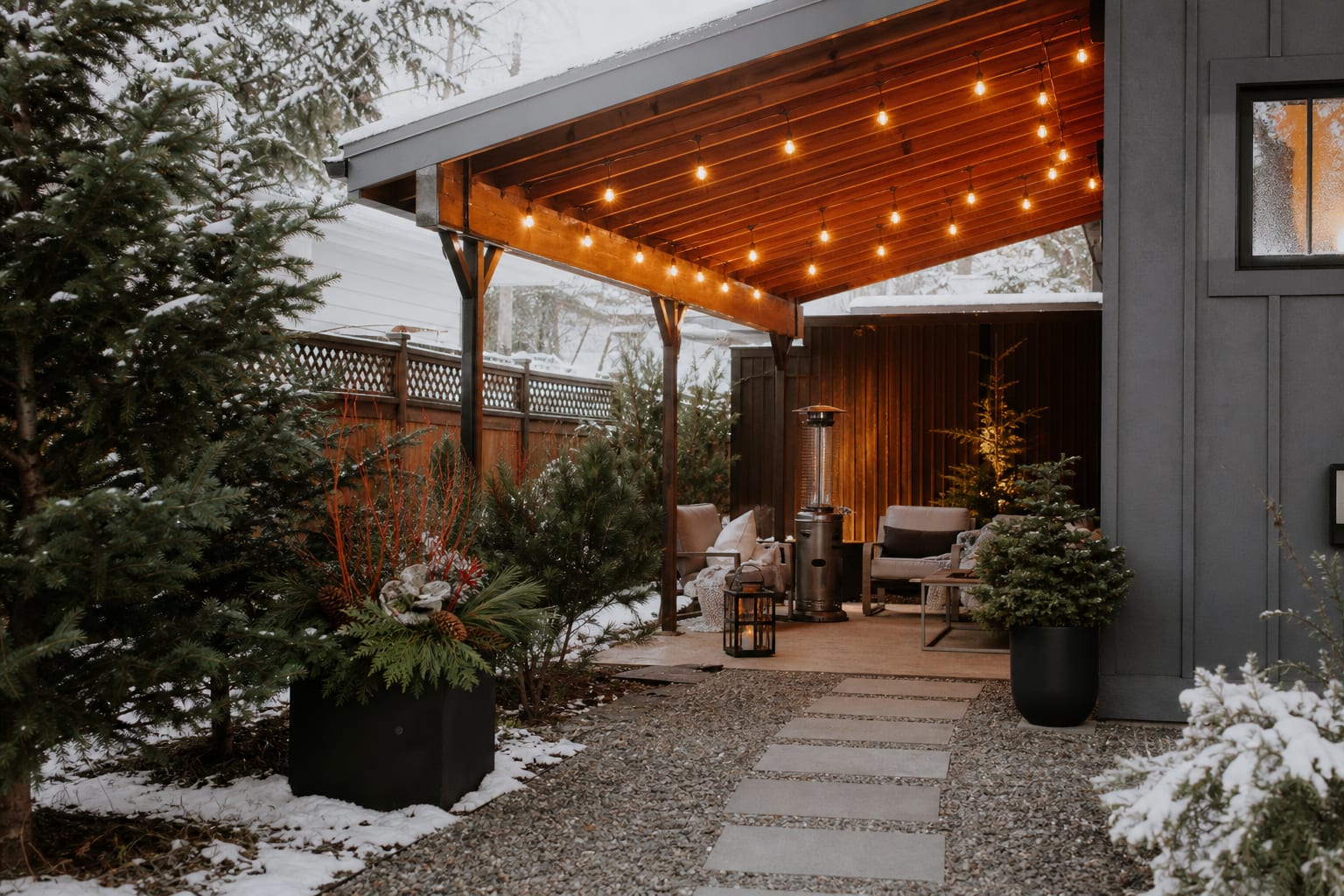 winter backyard with covered seating, warm string lights, evergreen planters, gravel path, and compact outdoor heater