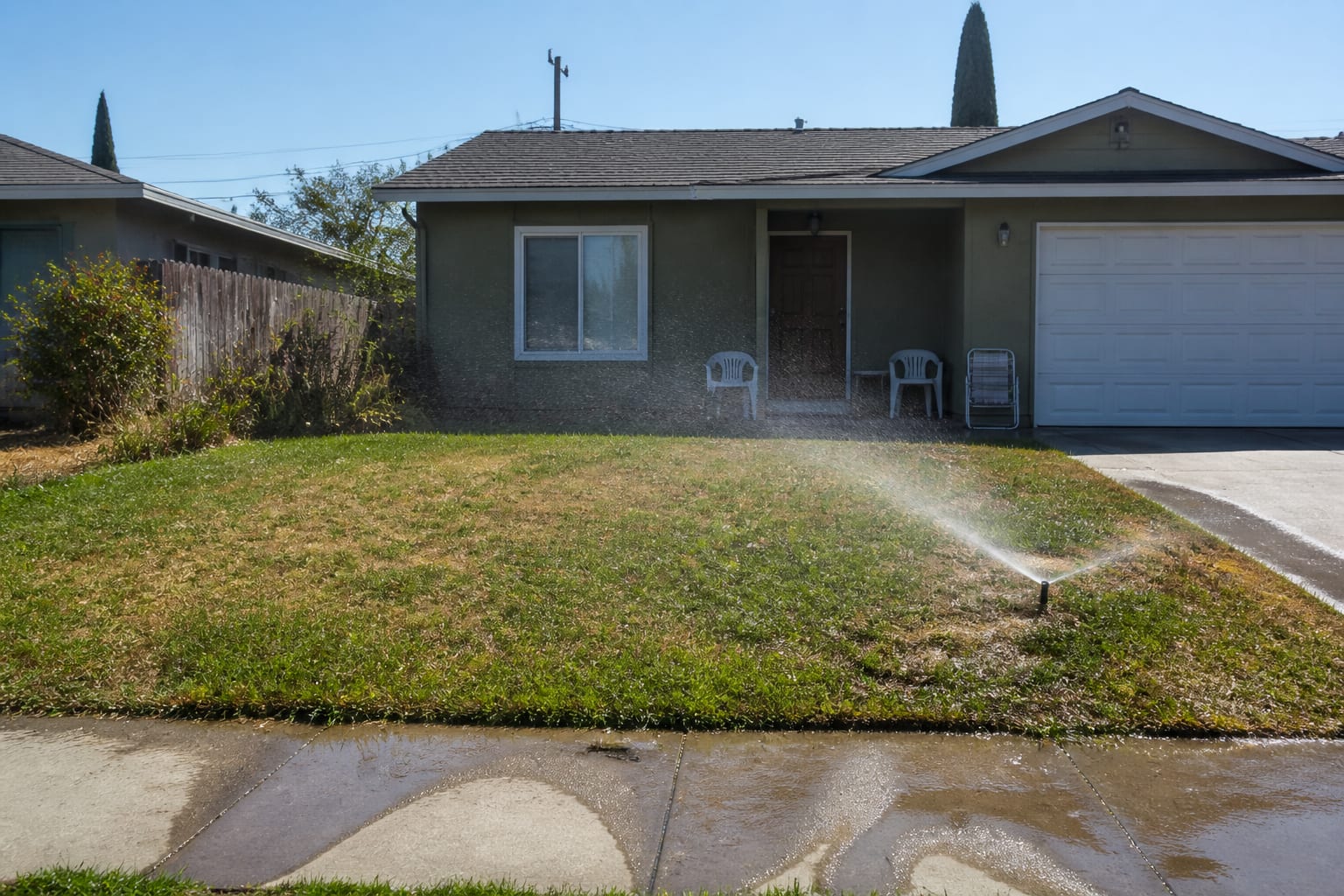 Thirsty patchy lawn with brown spots and a spray sprinkler running in midday sun before redesign
